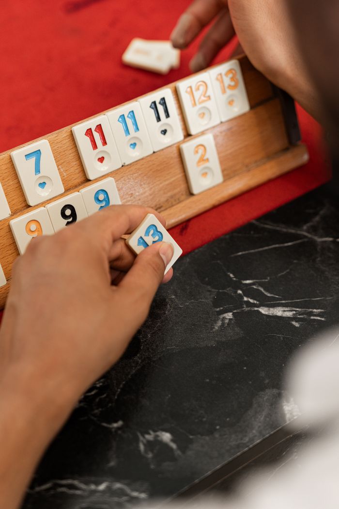 Close-up of a person arranging numbered tiles on a Rummikub board indoors, focusing on hands and tile numbers.